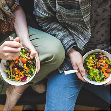 Two people sitting next to each other each holding a bowl with salad